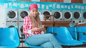 Young woman using smartphone while waiting for laundry, sitting on blue chairs in laundromat - Powered by Shutterstock - Get 15% off with code: PIKWIZARD15