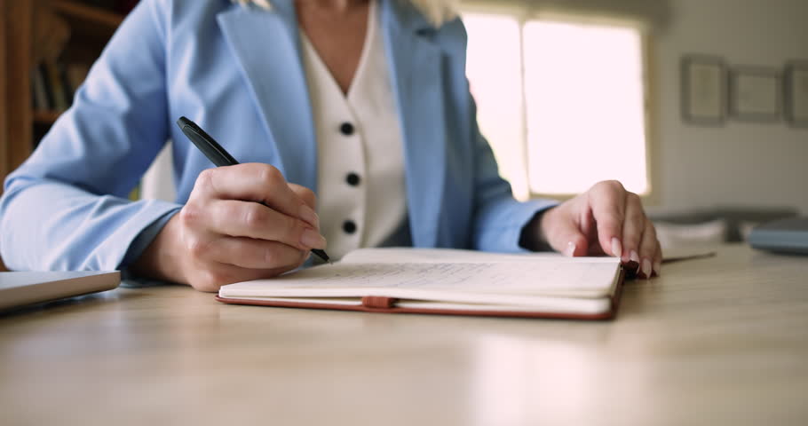 Professional woman writing in notebook seated at workplace desk, working or studying in office, carefully records information, busy in goals or to-do list creation, productivity, workflow organization