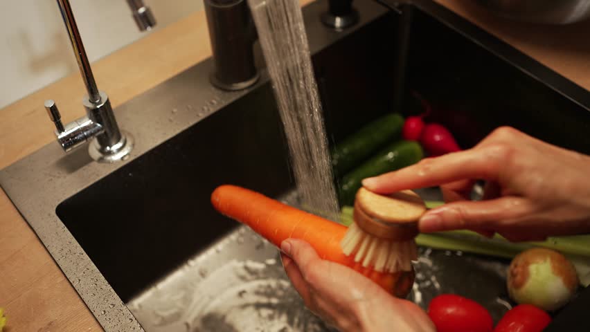 Chef washes a carrot with a small round brush under running water in a kitchen sink, with other fresh vegetables like celery, onions, tomatoes, and radishes visible in the background