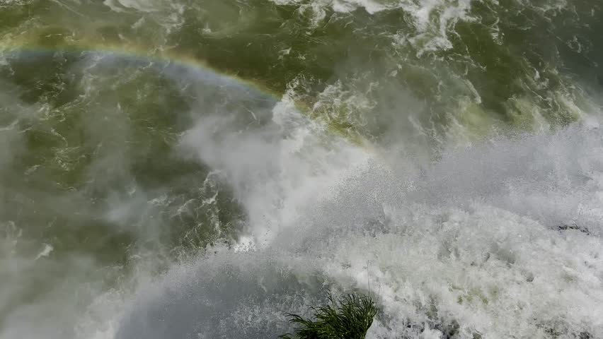 Rushing river waters with waterfalls and swirling mist reveal a vivid rainbow, creating a dramatic and colorful natural scene full of energy and wonder.