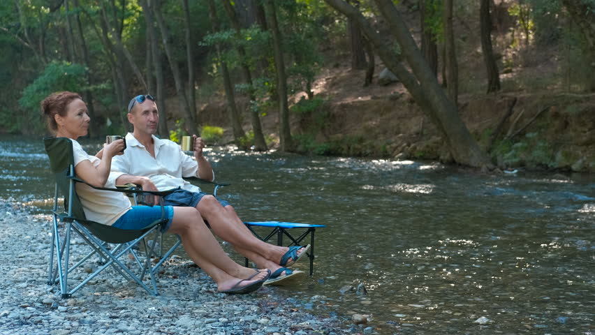 Couple enjoying coffee by the river in camping chairs. Peaceful riverside morning with tourists drinking coffee, resting in chairs and immersing themselves in serene natural surroundings