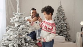 African American man films son decorating tree at home and saying hi, father takes smartphone photo of child at Christmas - Powered by Shutterstock - Get 15% off with code: PIKWIZARD15