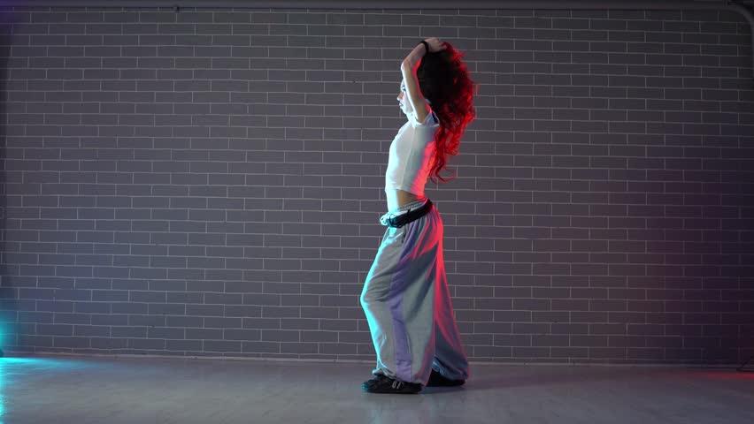 young crazy girl dancing hip hop against wall background in neon lighting, female choreographer moving to music and performing jazz funk in dance studio