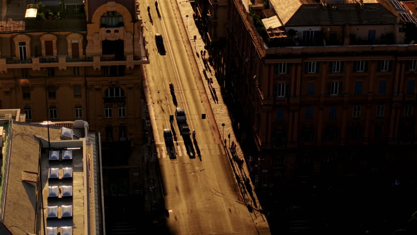 Low drone flight between buildings revealing a sunlit street in Genoa with vehicles and long shadows at sunset