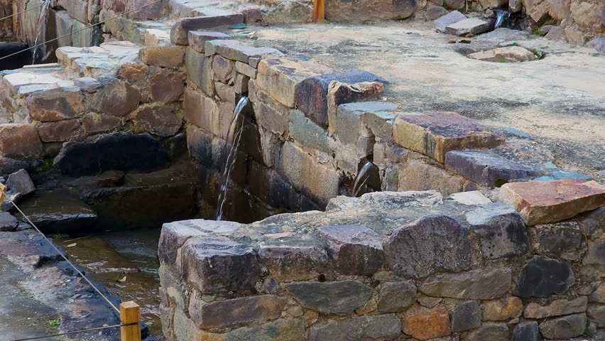 A cinematic pan shot captures the ancient Inca water fountains and intricate stone channels of the Ollantaytambo Archaeological Park, showcasing their historic and cultural significance.