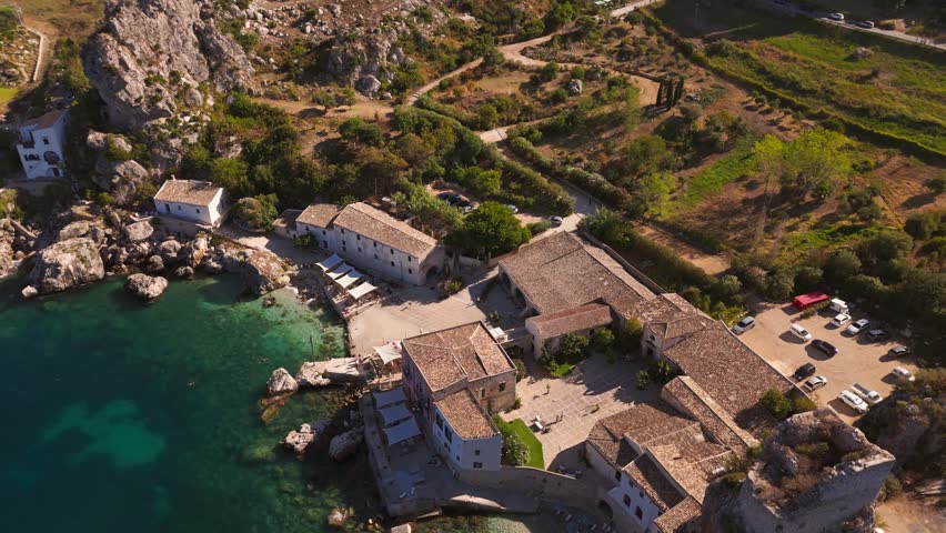 Aerial drone shot panning left capturing tourists on private beach and the sunlit blue ocean shining brightly at Tonnara di Scopello, Sicily, Italy, during summer