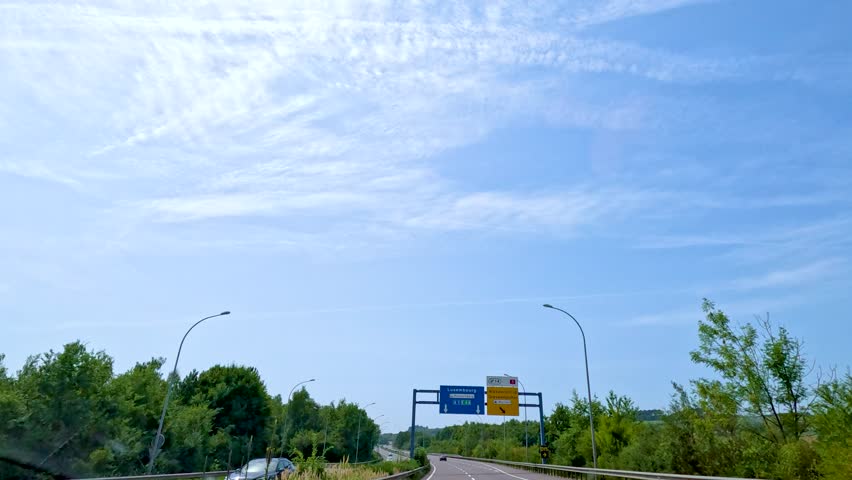 A vehicle travels along a tree-lined highway in Luxembourg, passing directional road signs under bright daylight with clear blue skies and scattered clouds
