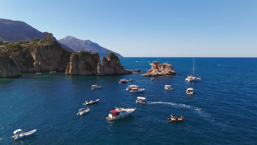Aerial drone footage orbiting right around the historic Tonnara di Scopello, showcasing the rugged Sicilian rocky coastline and a boat on the sparkling blue ocean in Italian summer in Sicily, Italy