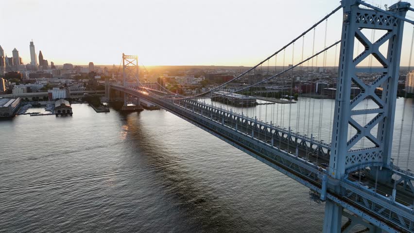 Iconic Benjamin Franklin Streep bridge, piers and skyline at golden sunrise. Philadelphia downtown in Pennsylvania. Aerial orbit wide shot. Cars crossing river from Camden.