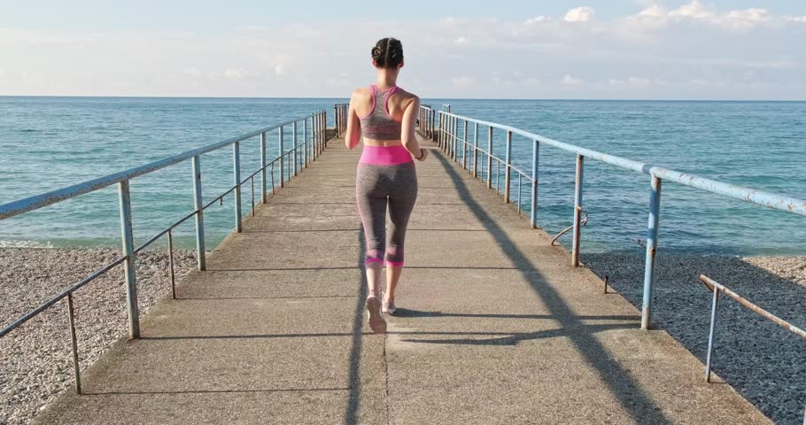 Woman jogging along concrete pier towards open sea morning sunlight, slow motion capturing fitness, freedom and coastal workout energy. Effect of blocked stabilization. LOCKED-ON STABILIZATION EFFECT