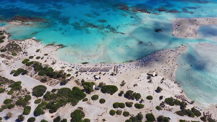 Aerial view of Elafonisi Beach in Crete, showing turquoise waters, sandbanks, rocky islets, and smooth camera motion over the serene coastline.