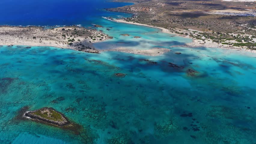 Smooth aerial pan of Elafonisi Beach in Crete, Greece, showing turquoise waters, white sandy shores, islets, lagoons, and distant mountains.