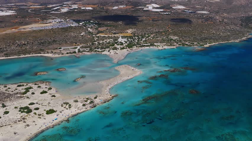 Aerial panning view of Elafonisi Beach in Crete, Greece, showing turquoise waters, sandy shores, rocky islets, and surrounding rugged coastline.