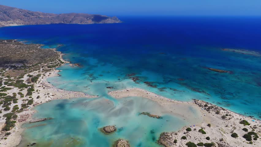 Smooth aerial pan over Elafonisi Beach in Crete, Greece, showing pinkish sands, turquoise waters, rocky islets, and rugged mountains along the coast.