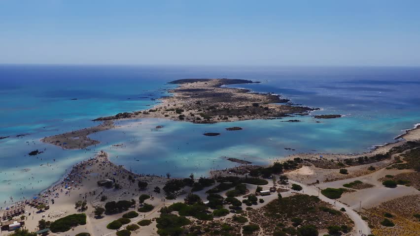 Aerial shot of Elafonisi Beach in Crete, showing pink sand, turquoise waters, shallow lagoons, and visitors wading near a narrow land strip.