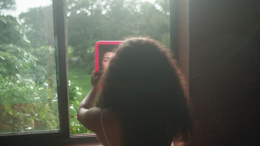Fixed shot of woman from behind holding a red mirror at a window, reflecting her face with garden greenery outside