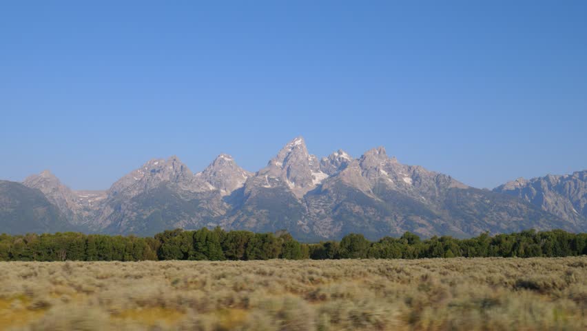 Majestic peaks of the Teton Range rise above forested slopes and golden grasslands in Grand Teton National Park. A crisp, clear view of Wyoming’s rugged alpine beauty under blue skies.