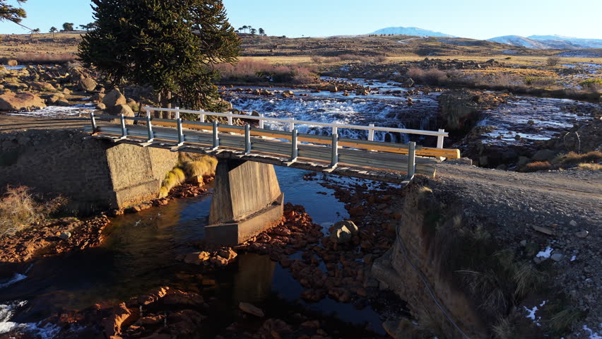 Elevated view of a small wooden bridge crossing the Rio Agrio above gentle cascades, framed by Andean steppe and volcanic terrain near Caviahue, Neuquén, in soft morning light.