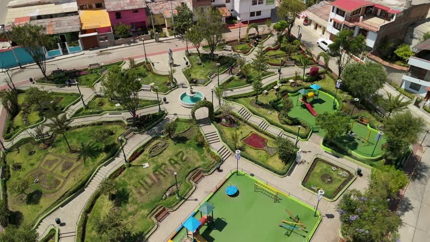 A dynamic aerial parallax shot circles over the beautifully landscaped Parque Santiago in Huaraz, Peru, revealing its intricate paths, fountain, playground, and surrounding city buildings.