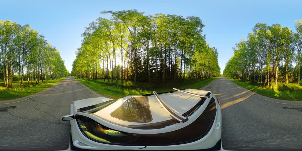 360 VR panorama of a white car moving along the green alley on an empty paved road during sunset with sun blinking through trees