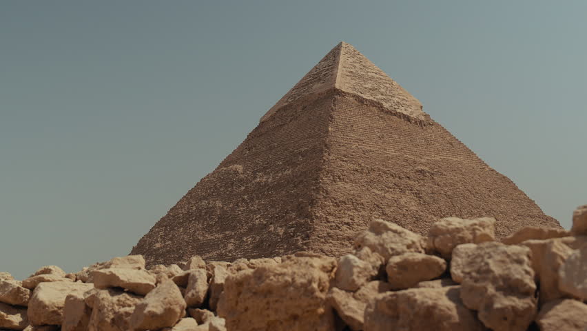 Wide shot of the Great Pyramid of Giza in Egypt on a sunny clear day. The pyramid stands tall with sandy rocks in the foreground, blue sky above, and desert scenery around this famous world landmark.