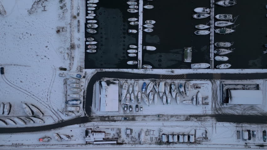 This aerial drone footage offers a top-down view of a snow-covered marina in winter, where dozens of boats float in dark, icy waters while others are neatly lined up on land