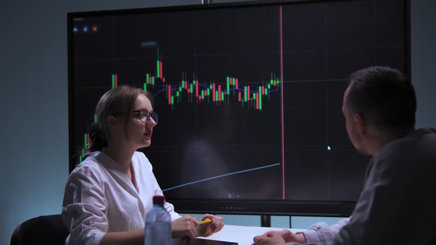 Woman and man at table in high-tech office analyzing stock market charts on screen