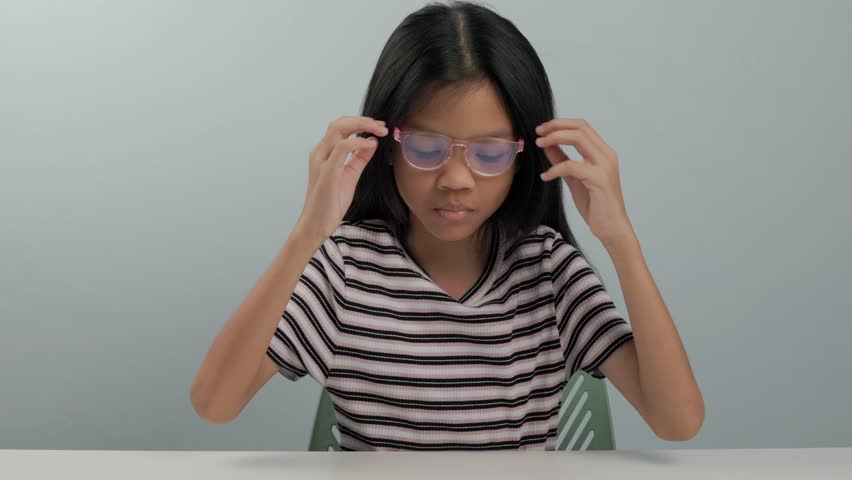 A kid primary girl is wearing glasses to block blue light from a screen. She removes her glasses and rubs her left eye because it feels irritated, itchy, and uncomfortable. The background is plain.