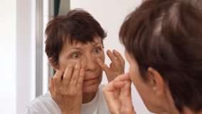 Mature woman examining puffiness and under-eye bags in the mirror. Natural signs of aging, skincare awareness, and self-care moment. Age-related skin changes, fatigue. Cosmetology and beauty concept - Powered by Shutterstock - Get 15% off with code: PIKWIZARD15