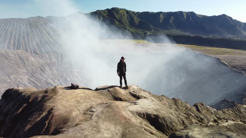 Smooth drone shot of a man standing at the edge of Mount Bromo’s smoking crater. The camera pulls back and rises, revealing the immense volcanic landscape in cinematic 4K.