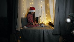 Cute child in Santa hat sitting by window with fairy lights and Christmas decorations, writing in notebook. Cozy winter holiday atmosphere at home. High quality 4k footage - Powered by Shutterstock - Get 15% off with code: PIKWIZARD15