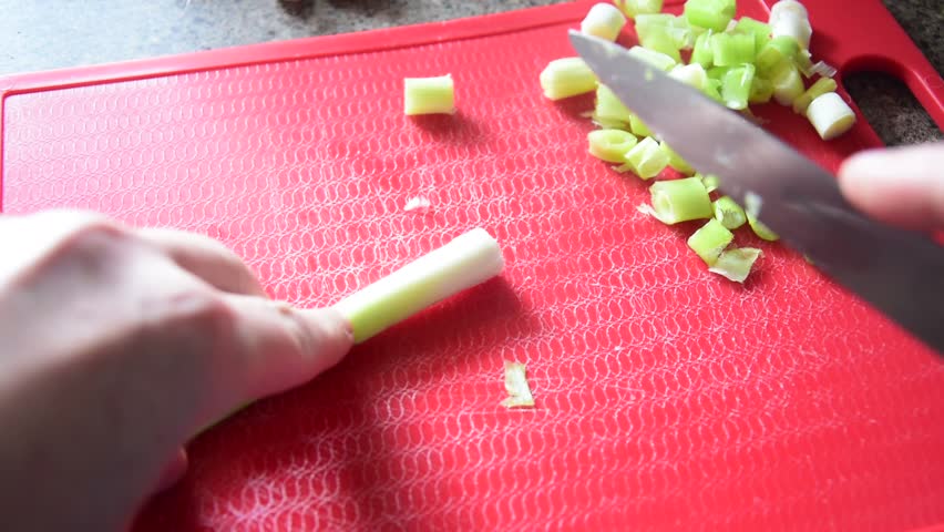 Close-up of hands chopping spring onions on a red chopping board with a silver knife, sunlight illuminating the board. Perfect for cooking, healthy food, recipe tutorials, or kitchen content.