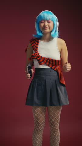 Cheerful blue-haired Asian woman dancing in headset on crimson background, k-pop
