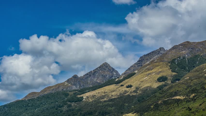 Sharp alpine peaks and forested hills near Glenorchy, New Zealand, under a blue sky with dramatic cumulus clouds in summer.