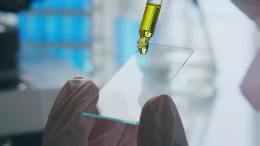 Close up view of gloved hands of scientist applying oil sample to glass slide with pipette for microscopic examination in laboratory