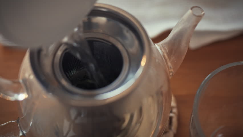 Hot water is being poured into a silver teapot that contains loose leaf tea. This takes place in a cozy kitchen setting, perfect for an afternoon tea experience