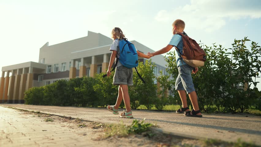 Child walking to school holding hand with backpack and friend near campus building under warm sunlight showing boy girl bond and casual step toward entrance during summer on paved walkway near hedge