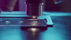 Closeup of microscope objective aligned above slide with evenly spaced droplets reflecting colorful lab lighting during precise scientific investigation - Powered by Shutterstock - Get 15% off with code: PIKWIZARD15