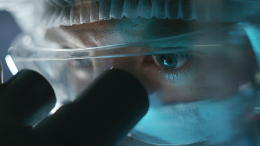 Concentrated scientist wearing face mask and safety glasses observing sample through microscope eyepiece in laboratory. Detailed close-up view