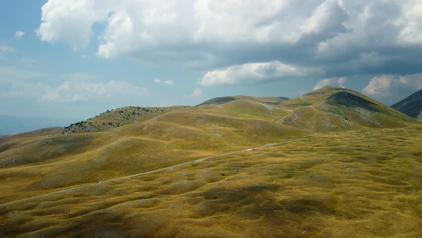 4K aerial drone footage flying forward over yellow and green grassy hills with single road and car at the base, distant mountain ranges in background.