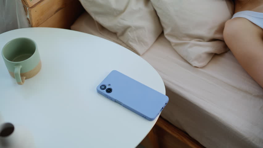Tired woman looking alarm smartphone lying at home bed closeup. Upset brunette taking mobile phone covering soft blanket in light bedroom. Sleepless young lady waking up in apartment early morning.