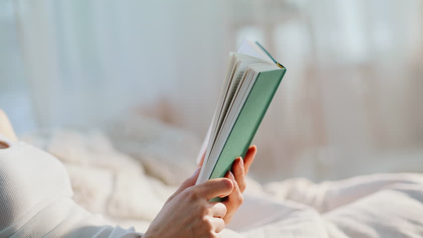 Woman hands holding book in white bedroom interior closeup. Smiling beautiful lady flipping pages enjoying literature hobby early morning. Happy reader drinking coffee reading romantic novel in bed.