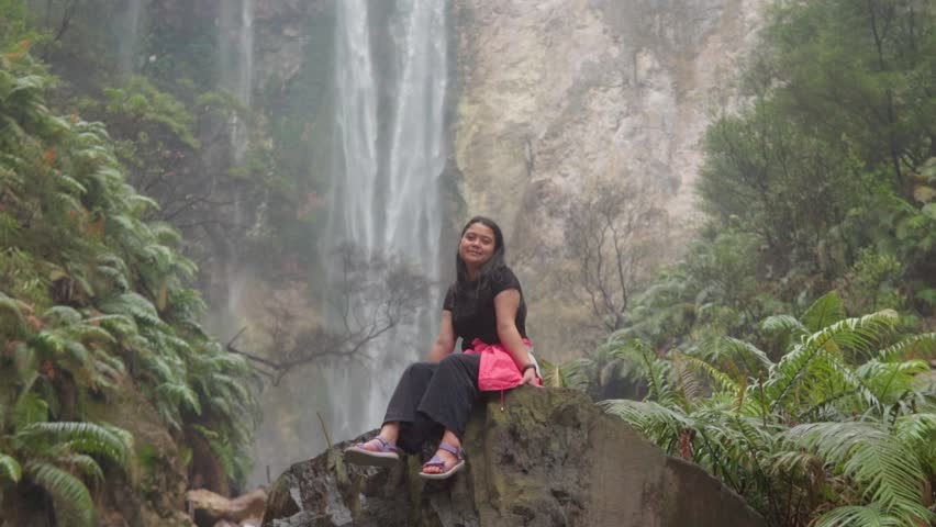 Slow motion video of an Asian woman sitting on a rock near a waterfall in a lush tropical forest, showing natural beauty, travel lifestyle, and outdoor adventure atmosphere.