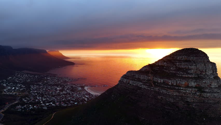 Cinematic aerial view of Table Mountain in Cape Town, South Africa, dramatic rocky cliffs with coastal cityscape at sunset, clouds rolling over peaks and golden evening light