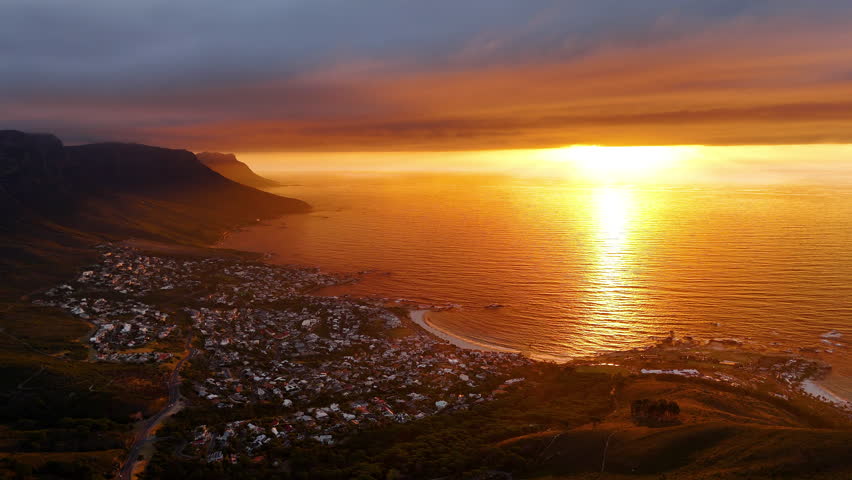 Cinematic aerial view of Table Mountain in Cape Town, South Africa, dramatic rocky cliffs with coastal cityscape at sunset, clouds rolling over peaks and golden evening light