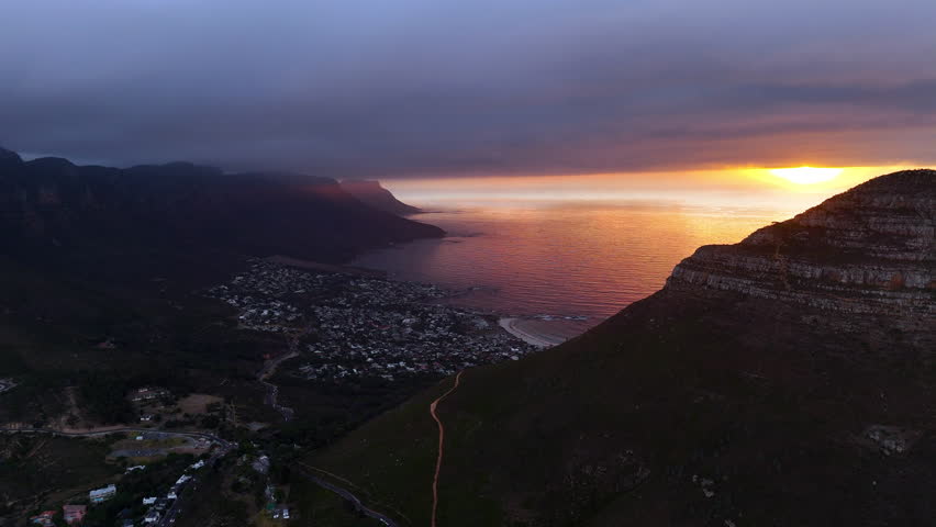 Cinematic aerial view of Table Mountain in Cape Town, South Africa, dramatic rocky cliffs with coastal cityscape at sunset, clouds rolling over peaks and golden evening light