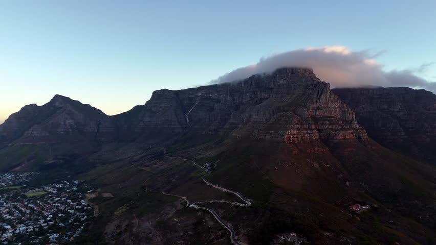 Cinematic aerial view of Table Mountain in Cape Town, South Africa, dramatic rocky cliffs with coastal cityscape at sunset, clouds rolling over peaks and golden evening light