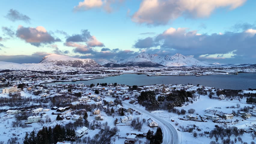 Aerial view of winter town in Lofoten, Norway, surrounded by snow-covered houses, mountains, and dramatic arctic landscape under soft evening light
