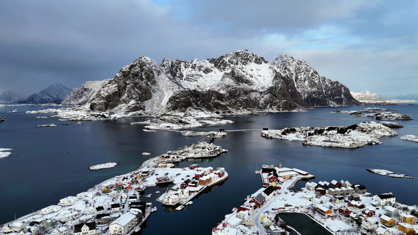 Cinematic aerial view of winter town in Lofoten, Norway, surrounded by snow-covered houses, mountains, and dramatic arctic landscape under soft evening light
