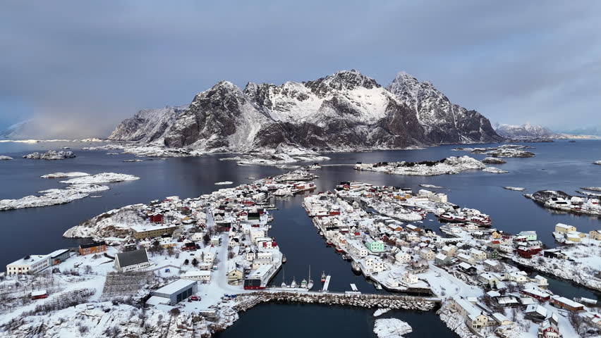 Cinematic aerial view of snow-covered mountains in Lofoten, Norway, dramatic winter landscape with icy valley, scattered houses and evening light over rugged arctic peaks